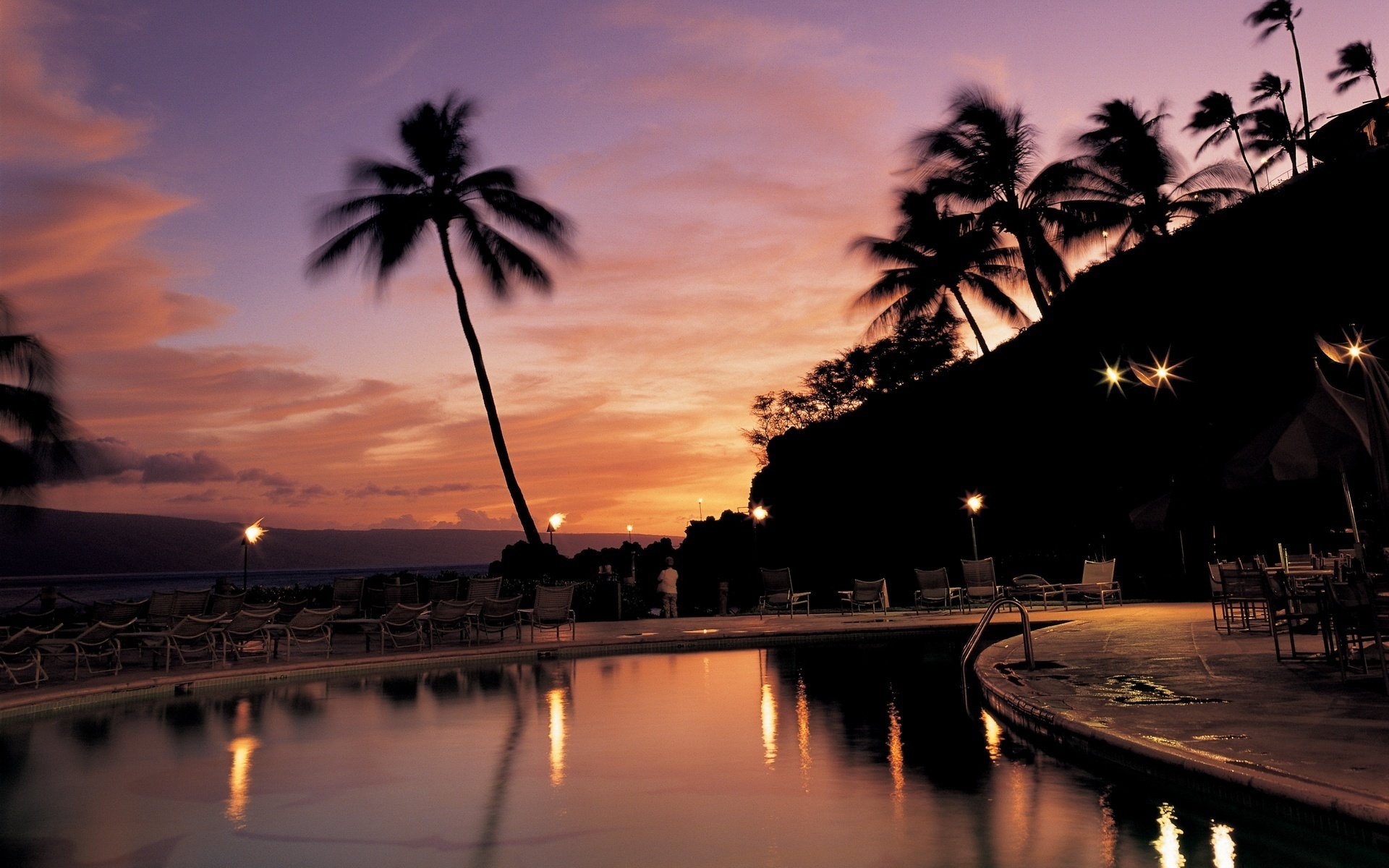 plan d eau soirée lanternes chaises longues piscine coucher de soleil palmiers ciel loisirs hôtel station balnéaire chaises longues silhouettes pénombre arbres tropiques ciel rose lumières réflexion